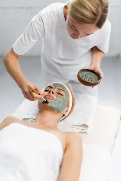 Blonde Kosmetikerin trägt Tonmaske auf Gesicht einer Frau auf, die auf Massagetisch liegt — Stockfoto