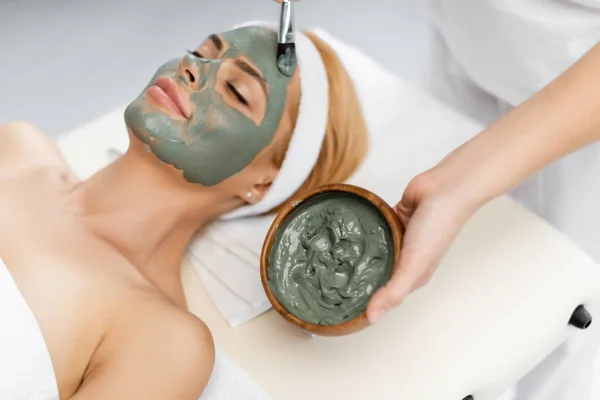 Vista de ángulo alto de esteticista aplicación de máscara de arcilla en la cara de la mujer acostada en la mesa de masaje - foto de stock
