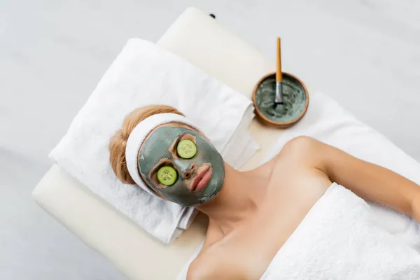 Draufsicht der Frau mit Lehmmaske und Gurkenscheiben auf Augen, die auf Massagetisch liegen — Stockfoto