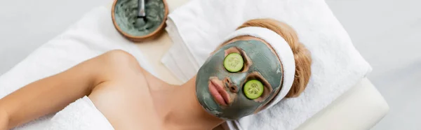 Draufsicht auf Frau mit Lehmmaske und Gurkenscheiben auf Augen, die auf Massagetisch liegen, Banner — Stockfoto