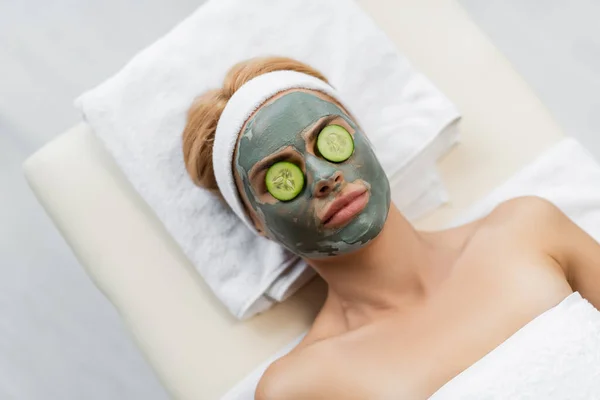 Draufsicht der Frau mit Lehmmaske und frischen Gurkenscheiben auf Augen, die auf Massagetisch liegen — Stockfoto