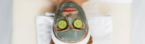 Vista dall'alto della donna con maschera di argilla e fette di cetriolo fresco sugli occhi sdraiati sul tavolo da massaggio, banner — Foto stock