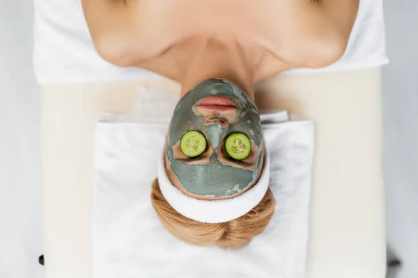Vista superior de la mujer con máscara de arcilla y en rodajas de pepino fresco en los ojos acostado en la mesa de masaje - foto de stock