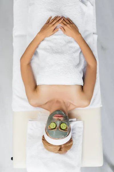 Top view of blonde woman with clay mask and sliced fresh cucumber on eyes lying on massage table — Stock Photo