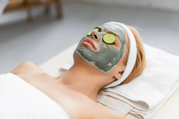 Femme blonde avec masque en argile et tranches de concombre sur les yeux couchés sur la table de massage — Photo de stock