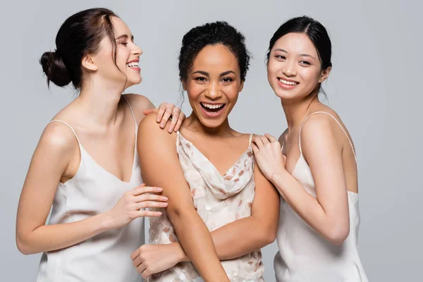 Позитивні багатоетнічні жінки в шовкових сукнях позують ізольовані на сірому — стокове фото