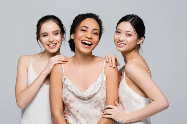 Smiling multiethnic women posing and looking at camera isolated on grey — Stock Photo