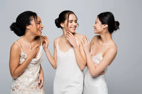 Felices mujeres multiétnicas en vestidos de seda hablando aisladas en gris - foto de stock