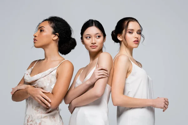 Jóvenes mujeres interracial en vestidos de satén posando aislados en gris - foto de stock