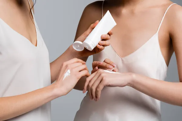 Vista recortada de la mujer en vestido de seda aplicando crema en la mano cerca de amigo aislado en gris - foto de stock