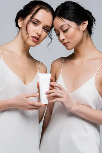 Young multiethnic women in silk dresses holding cosmetic cream isolated on grey — Stock Photo