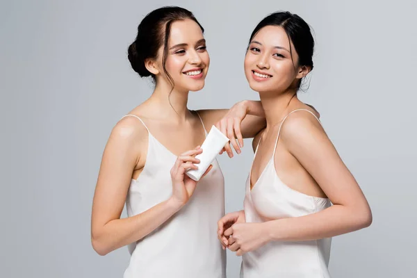 Heureuses femmes multiethniques en satin tenant la crème en tube isolé sur gris — Photo de stock