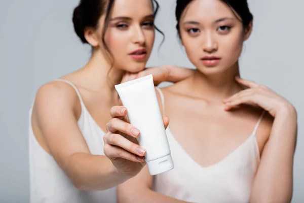 Mulher desfocada segurando creme cosmético perto de asiático amigo isolado em cinza — Fotografia de Stock