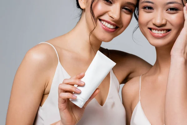 Усміхнена жінка тримає косметичний крем біля азіатського друга ізольовано на сірому — стокове фото