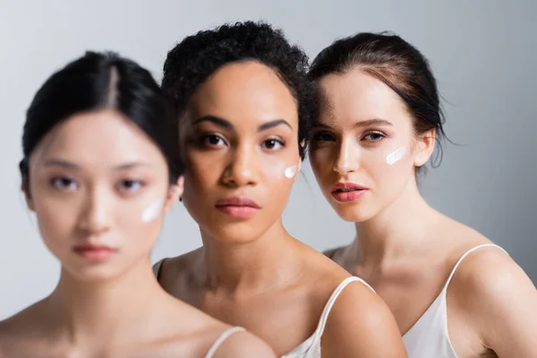 Young woman with cream on face looking at camera near blurred interracial friends isolated on grey — Stock Photo