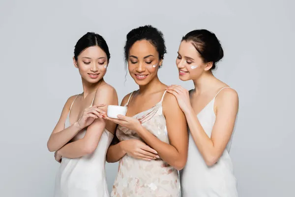 Mujeres bastante interracial en vestidos de satén que sostienen crema cosmética aislada en gris — Stock Photo