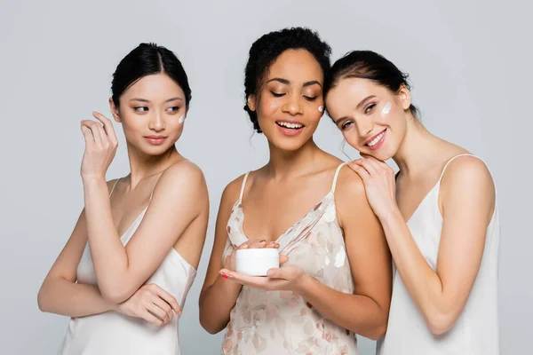 Mujer afroamericana sosteniendo crema cosmética cerca de amigos interracial aislados en gris — Stock Photo
