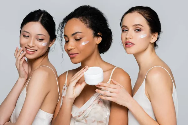 Mujer afroamericana sosteniendo crema cosmética cerca de amigos multiétnicos posando aislados en gris — Stock Photo