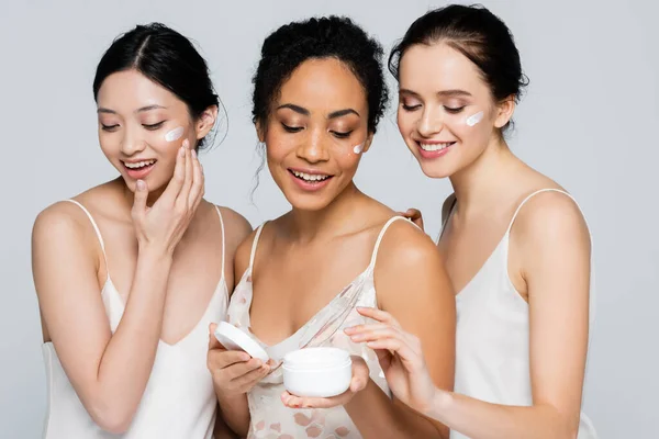 Happy multicultural women applying cosmetic cream on faces isolated on grey — Stock Photo