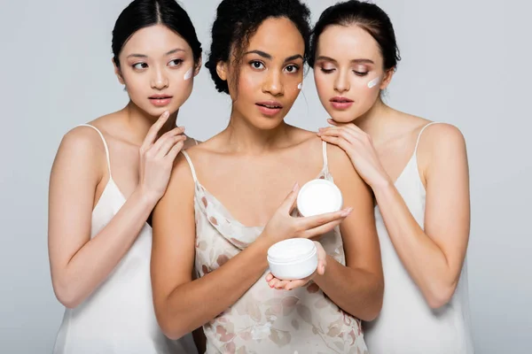African american woman with cosmetic cream looking at camera near multicultural friends isolated on grey — Stock Photo