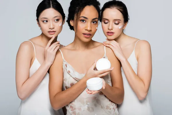 African american woman with cosmetic cream looking at camera near multicultural friends isolated on grey — Stock Photo