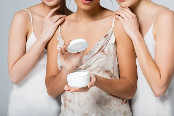 Vista cortada de jovens mulheres inter-raciais com creme cosmético isolado em cinza — Fotografia de Stock