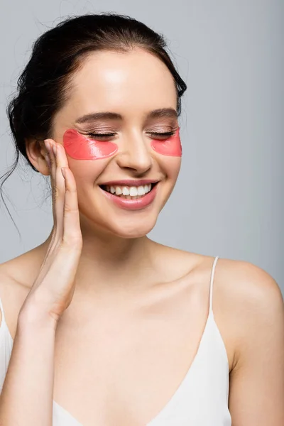 Femme gaie avec des patchs pour les yeux touchant le visage isolé sur gris — Photo de stock