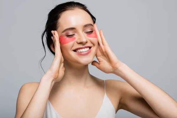 Femme souriante avec des patchs oculaires touchant le visage isolé sur gris — Photo de stock