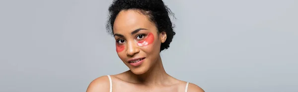 Mujer afroamericana con parches en los ojos sonriendo a la cámara aislada en gris, pancarta - foto de stock