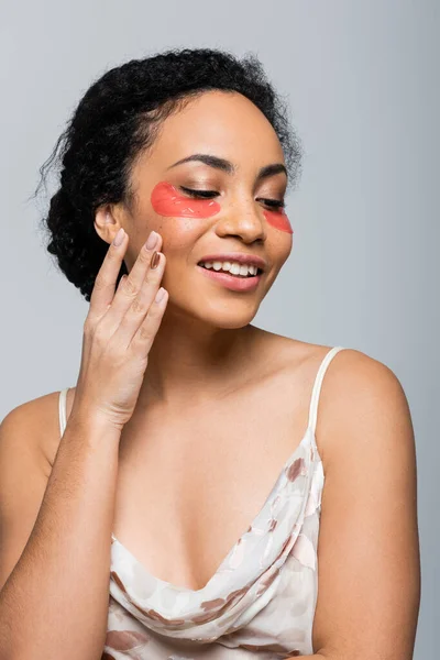 Sorridente donna afro-americana in bendaggi oculari toccando guancia isolata su grigio — Foto stock