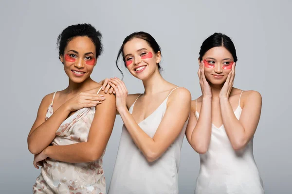 Sonrientes mujeres multiétnicas con parches en los ojos mirando a la cámara aislada en gris - foto de stock