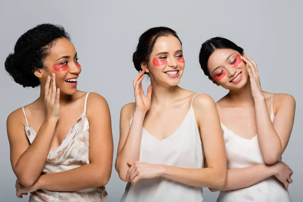 Hübsche interrassische Frauen mit Augenklappen, die Gesichter berühren, isoliert auf grau — Stockfoto