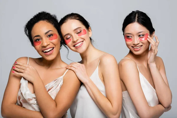 Alegres amigos multiculturales en parches para los ojos y vestidos de satén posando aislados en gris - foto de stock