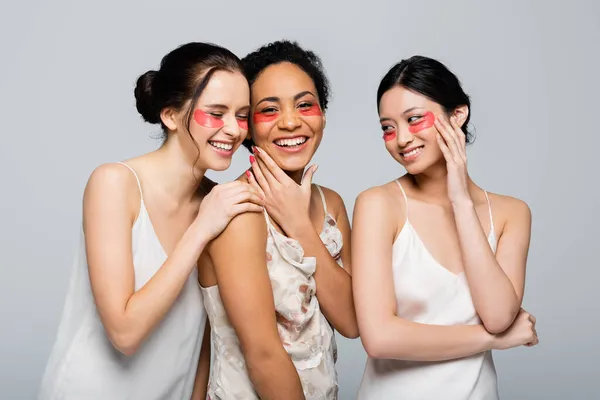 Mujeres alegres multiculturales en vestidos de seda y parches para los ojos aislados en gris - foto de stock