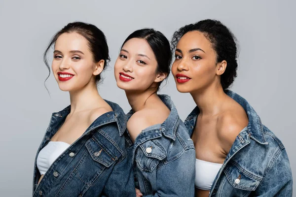 Lächelnde multikulturelle Frauen mit roten Lippen, die isoliert auf grau in die Kamera schauen — Stockfoto