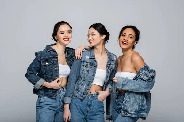 Pretty multiethnic women with red lips and denim jackets looking at camera isolated on grey — Stock Photo