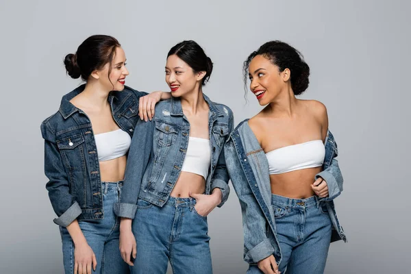 Giovane donna guardando sorridenti amici interrazziale in giubbotti di jeans isolati su grigio — Foto stock