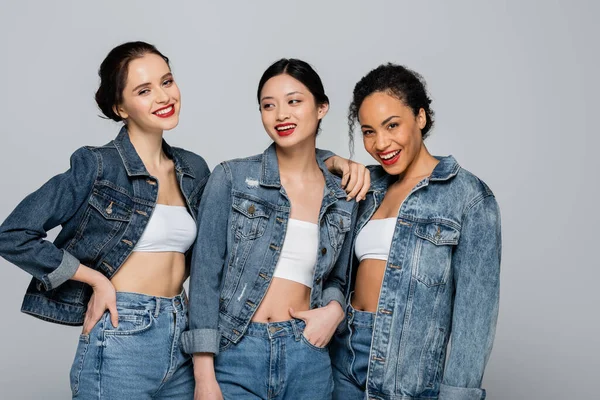 Sonrientes mujeres interracial con labios rojos y chaquetas de mezclilla posando aisladas en gris — Stock Photo