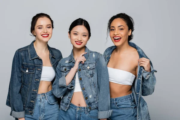 Mujer asiática con labios rojos sonriendo cerca de amigos interracial en chaquetas de mezclilla aisladas en gris - foto de stock