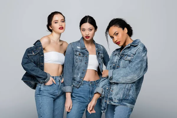 Pretty interracial women in denim jackets looking at camera isolated on grey — Stock Photo