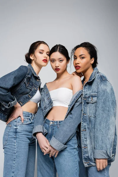 Low angle view of young interracial women in denim jackets looking at camera isolated on grey — Stock Photo