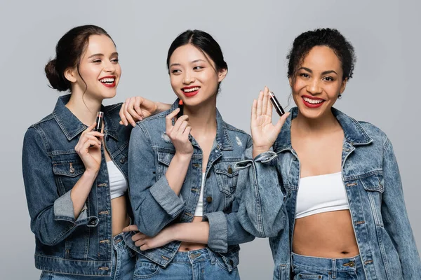 Lächelnde multiethnische Frauen in Jeansjacken, die Lippenstifte isoliert auf grau halten — Stockfoto