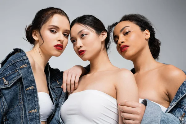 Low angle view of interracial women with red lips looking at camera near asian friend isolated on grey — Stock Photo