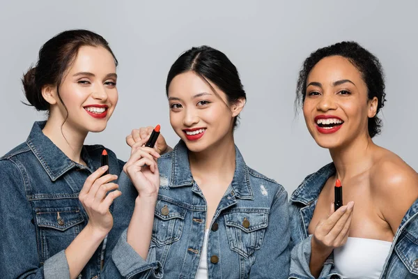 Smiling interracial friends holding red lipsticks isolated on grey — Stock Photo