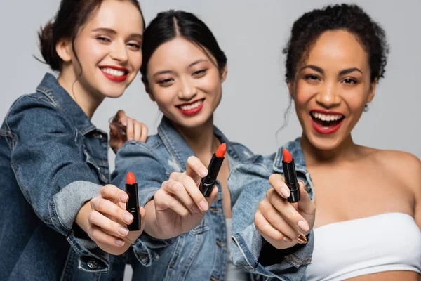 Lápiz labial rojo en manos de alegres mujeres interracial sobre fondo borroso aislado sobre gris - foto de stock