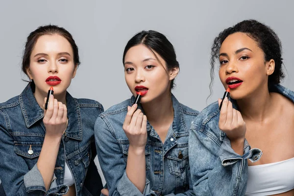 Hübsche multikulturelle Frauen tragen rote Lippenstifte auf und blicken vereinzelt in die Kamera auf grau — Stockfoto