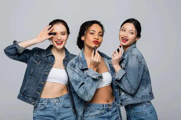 Young interracial women in denim jackets holding red lipsticks isolated on grey — Stock Photo