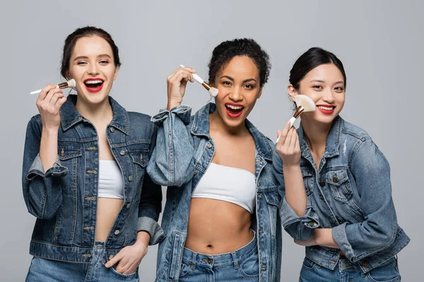 Glückliche multikulturelle Frauen in Jeansjacken, die Kosmetikbürsten isoliert auf grau halten — Stockfoto