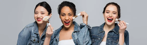 Fröhliche multiethnische Frauen in Jeansjacken mit Kosmetikbürsten auf grauem Banner — Stockfoto