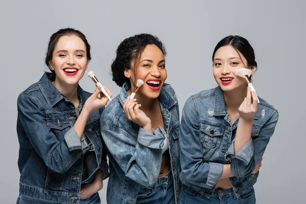 Happy multicultural women with red lipsticks holding cosmetic brushes isolated on grey — Stock Photo